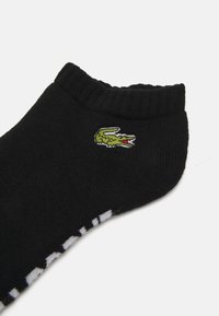 Chaussette noire en coton à la cheville avec une bordure côtelée. Présente un logo de crocodile brodé en vert et des lettres blanches le long du bas.