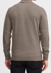 Golf beige lavorato a maglia con un collare alto, polsini e orlo a costine, con toppe sui gomiti e una piccola etichetta con logo vicino al fondo.