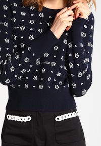 Marinblå tröja med vita hjärtan och fredssymboler, båda med ribbade muddar och nederkant. I kombination med svarta byxor med vita kedjeaccenter.