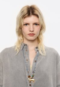 Collana dorata con pendente a forma di leone, con accenti di perline e un cordino blu, indossata su una camicia di denim grigia.
