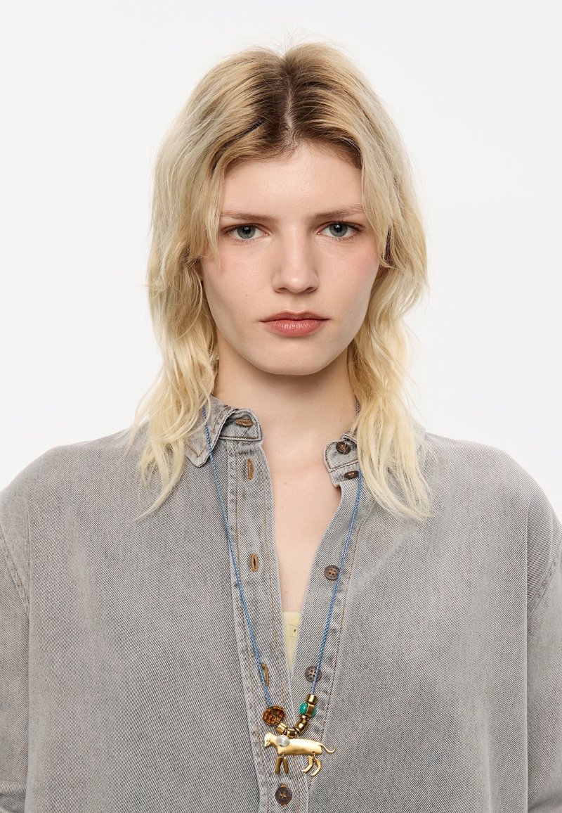 Collana dorata con pendente a forma di leone, con accenti di perline e un cordino blu, indossata su una camicia di denim grigia.