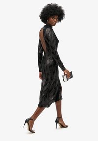 Vestido negro de lentejuelas con un patrón en zigzag, mangas largas, espalda descubierta y largo hasta la mitad de la pantorrilla. Combinado con tacones altos negros y un bolso metálico.