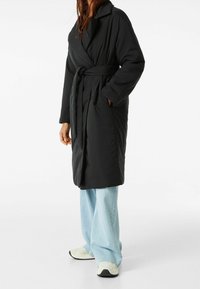 Svart vadderad wrap-kappa med bälte i midjan, bred krage, sidofickor och en slät yta, kombinerad med ljusblå vida jeans.