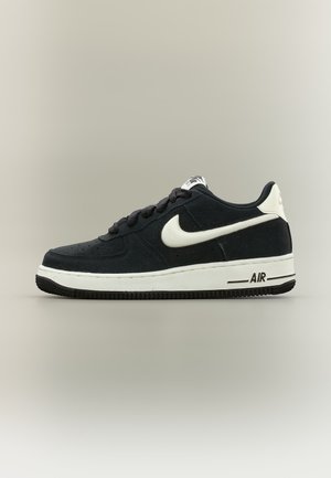 Baskets Nike Air Force 1 noir et blanc avec logo Swoosh blanc, languette de talon blanche et lacets noirs sur fond neutre.