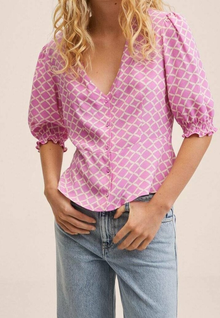 Femme portant un chemisier violet boutonné avec des motifs croisés blancs et des manches bouffantes, associé à un jean bleu clair.