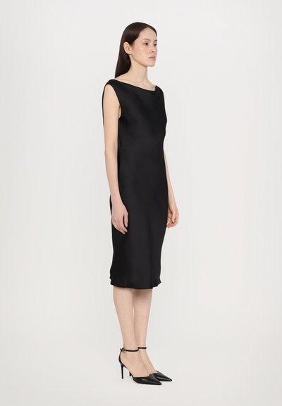 Vestido de satén negro con cuello barco, diseño sin mangas y abertura a la altura de la rodilla. Modelo lleva tacones negros de punta afilada con tiras en el tobillo.