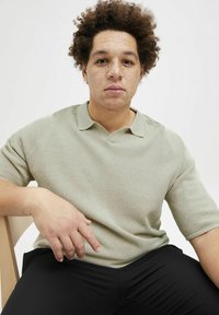 Polo shirt tricoté vert clair avec un col en V et des manches courtes, associé à un pantalon noir. Le tissu a une apparence texturée.