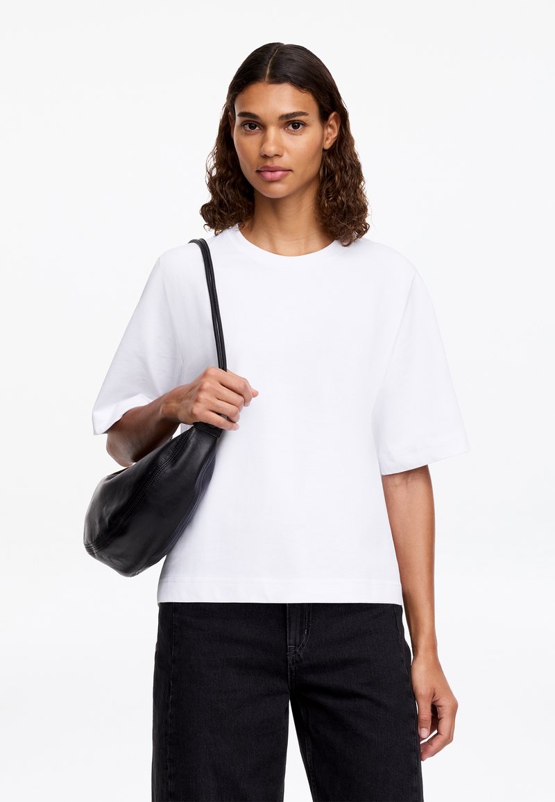 Femme aux cheveux bouclés porte une chemise blanche à manches courtes et un pantalon noir, portant un sac à épaule noir sur son épaule gauche sur un fond blanc.