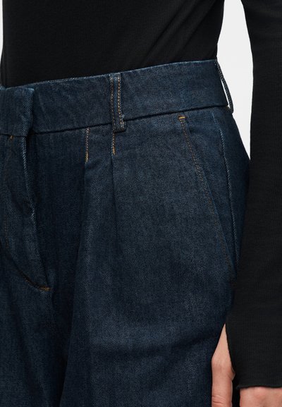 Σκούρα μπλε τζιν παντελόνια με πιέτες, φορεμένα από άτομο με μαύρο μακρυμάνικο τοπ, εστίαση στην περιοχή της μέσης και της τσέπης.