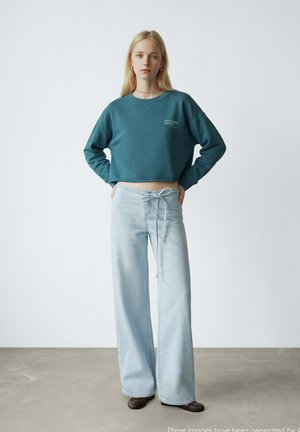 Giovane donna in piedi con le mani sui fianchi, indossa una felpa corta color teal e jeans larghi a gamba larga azzurri con vita a coulisse.