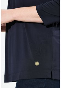 Tummansiniset lyhythihaiset paita, valmistettu sileästä kankaasta, ja siinä on pieni pyöreä kultainen yksityiskohta helman lähellä sekä sivuirrotus.