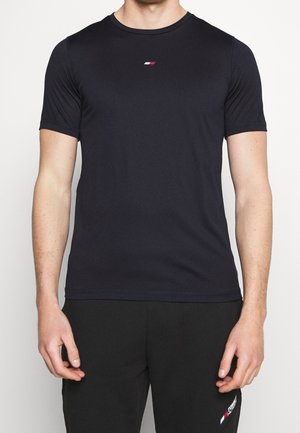 Mężczyzna ubrany w prostą czarną koszulkę z krótkim rękawem z małym logo w kolorach czerwonym, białym i niebieskim na środku klatki piersiowej oraz czarne spodnie.