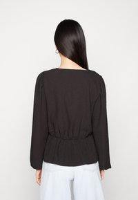 Blusa preta texturizada com mangas longas, apresentando um decote quadrado e uma cintura franzida. Usada com jeans azul claro e acessórios minimalistas.