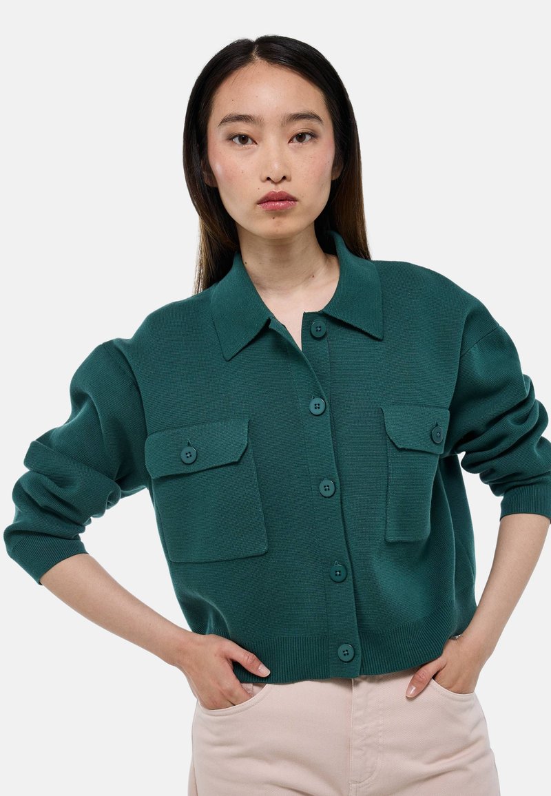 Cardigan en tricot bleu canard avec un col, deux poches poitrine et fermeture à boutons. Présente un ourlet et des poignets côtelés. Texture lisse.