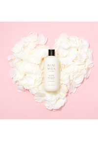 Bottiglia bianca di shampoo nutriente con tappo nero, etichettata "REAL SHEA," circondata da petali di rosa bianchi su uno sfondo rosa.