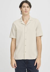 Camisa de manga corta beige con cuello en textura acanalada, con un frente abotonado y un ajuste relajado. Combinada con jeans oscuros.