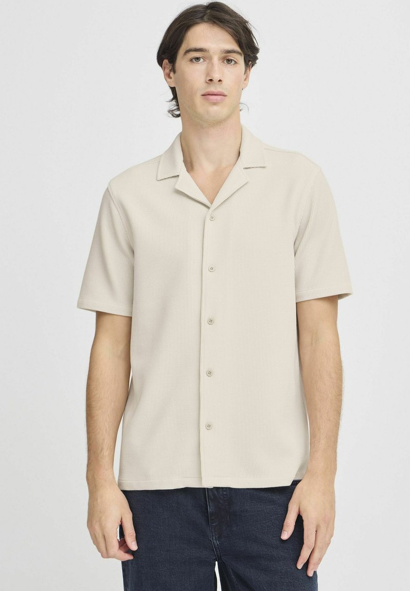 Camisa de manga corta beige con cuello en textura acanalada, con un frente abotonado y un ajuste relajado. Combinada con jeans oscuros.