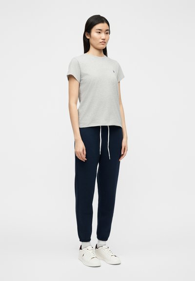 Νεαρή γυναίκα όρθια, φοράει ανοιχτό γκρι μπλουζάκι, σκούρα μπλε παντελόνια φόρμας με κορδόνια, λευκές κάλτσες και λευκά αθλητικά παπούτσια.