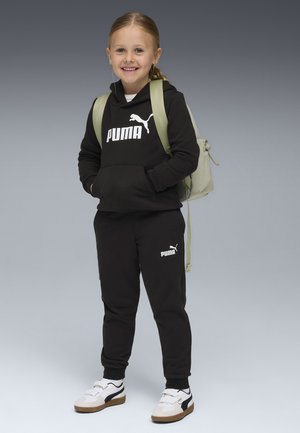 Haut à capuche noir et sweat-pants avec logo Puma blanc, associés à des chaussures blanches et marron. Sac à dos vert clair. Enfant souriant avec de longs cheveux.
