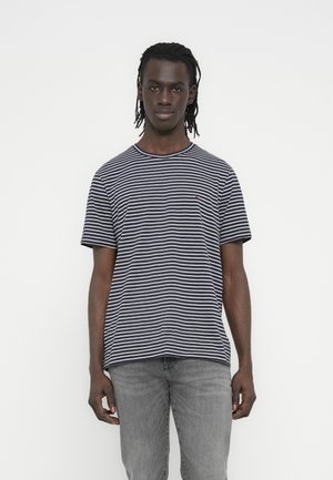 Młody mężczyzna o ciemnej karnacji z dredami, ubrany w czarno-biały pasiasty krótki rękaw koszuli i jasnoszare dżinsy, stojący na jednolitym białym tle.