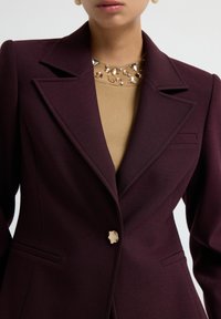 Femme portant un blazer ajusté violet foncé avec de larges revers sur un pull beige orné de perles en gemmes variées au niveau du col.