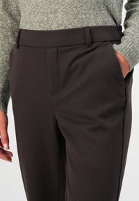 Pantalon sur mesure marron foncé avec une texture lisse, doté de poches latérales et d'une coupe avant plate. Associé à un pull en tricot vert clair.