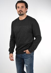 Fekete sweatshirt texturált első résszel, kerek nyakkal és bordás mandzsettákkal, elhalványult kék farmerral párosítva.