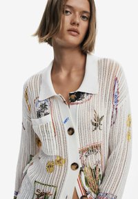 Baltas megztinis su aventūriniu mezgimu, puoštu spalvingais gėlių ir tropiniais raštais. Turi apykaklę, didelius medinius mygtukus ir kišenę.