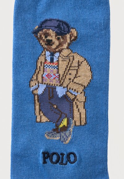 Ours en tricot portant une casquette plate, un pull, un manteau, un pantalon et des chaussures avec "POLO" brodé en dessous sur un tissu bleu.