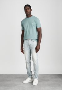 Lys grønn t-skjorte med korte ermer og liten logo, kombinert med falmede blå jeans og hvite sneakers, stående på en nøytral bakgrunn.