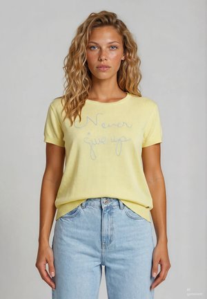 Jeune femme aux cheveux blonds ondulés portant un t-shirt jaune avec le texte « Never give up » et un jean taille haute bleu clair.