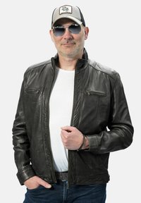 Zwarte leren jas met ritssluitingen, gebogen kraag en een gladde textuur. Gecombineerd met een wit shirt en blauwe jeans, accessoires zoals een zonnebril en een pet.