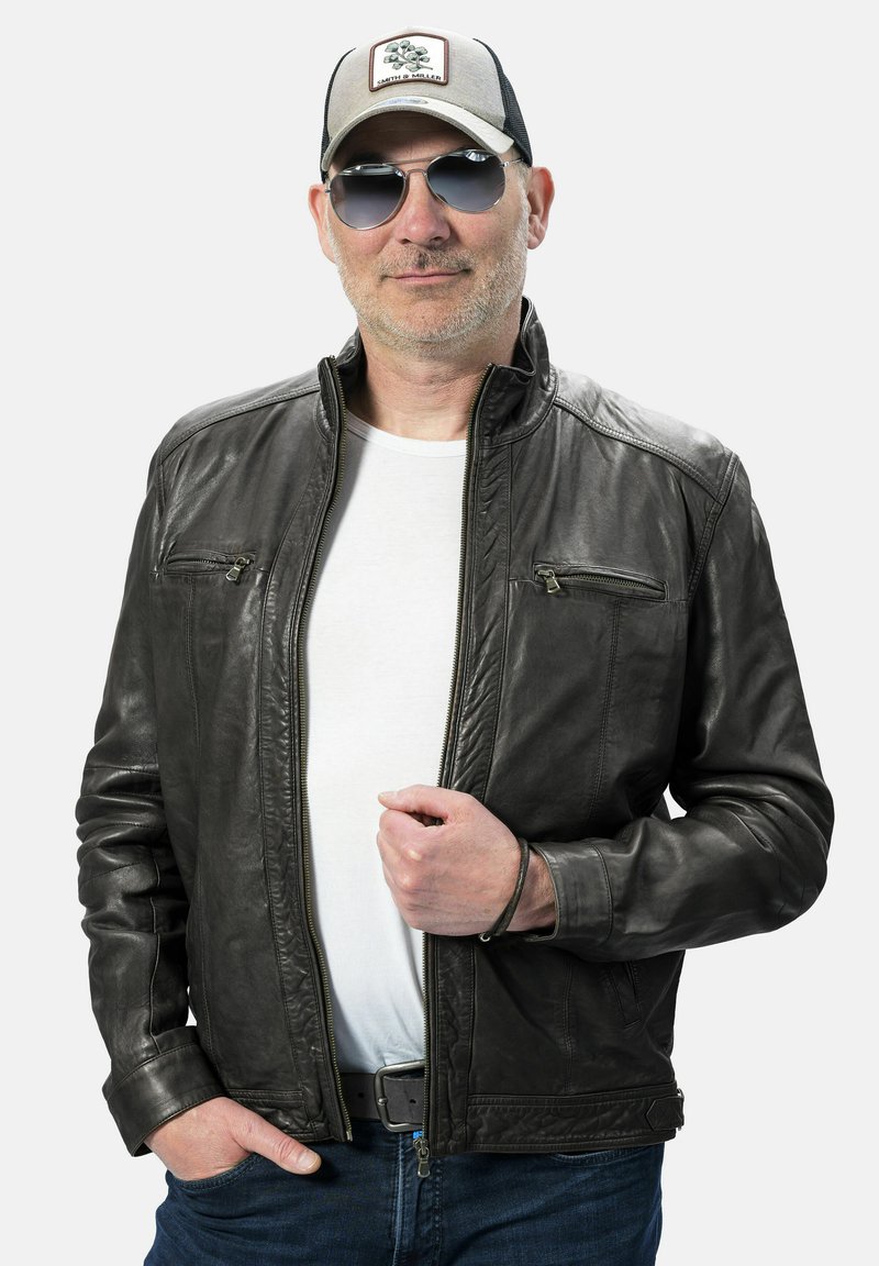 Zwarte leren jas met ritssluitingen, gebogen kraag en een gladde textuur. Gecombineerd met een wit shirt en blauwe jeans, accessoires zoals een zonnebril en een pet.