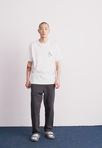 Weißes T-Shirt mit drei schwarzen grafischen Gesichtern auf der Brust, kombiniert mit dunkelgrauen Hosen und grauen Flip-Flops auf blauem Boden, Person steht.