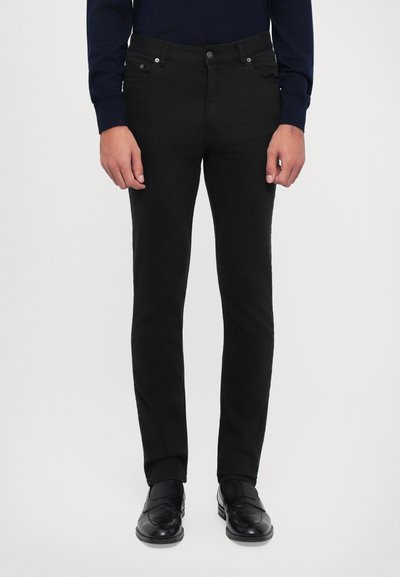 Schwarze Denim-Hose mit schmaler Passform, fünf Taschen, Metallbeschlägen und kontrastierenden Nähten. Getragen mit schwarzen Dressschuhen.