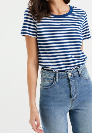 Vrouw draagt een blauw-wit horizontaal gestreept T-shirt met korte mouwen, ingestopt in een blauw spijkerbroek met hoge taille.