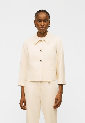 Femeie cu părul împletit, purtând o jachetă crem cu nasturi și guler, alături de pantaloni crem cu talie înaltă, pe un fundal simplu.