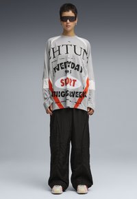 Grå langærmet skjorte med fed sort og orange grafisk tekst, parret med løse sorte bukser. Modellen bærer mørke solbriller og sneakers.