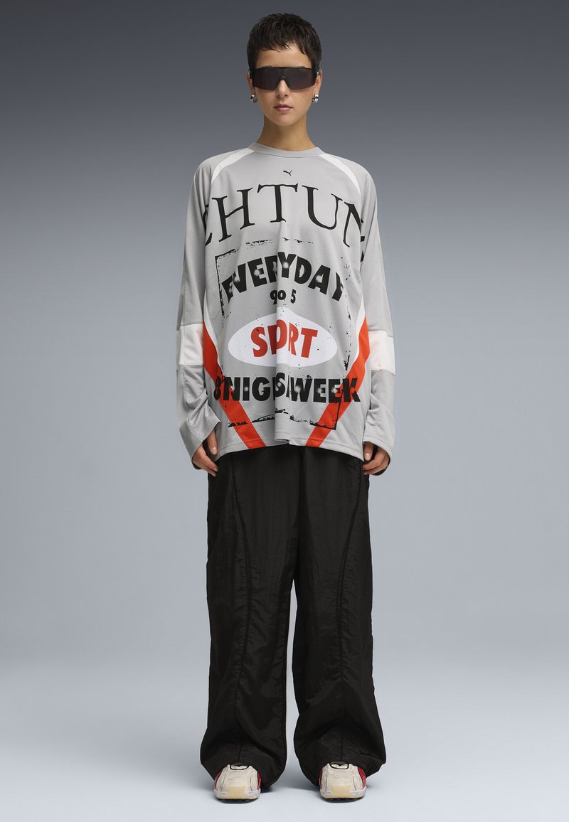 Grauhemd mit langen Ärmeln, das mit kräftigem schwarzen und orangefarbenem Grafiktext bedruckt ist, kombiniert mit lockeren schwarzen Hosen. Das Model trägt dunkle Sonnenbrillen und Sneakers.