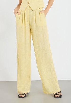 Kobieta ubrana w jasnożółte, wysokiego stanu spodnie o szerokich nogawkach z bocznymi kieszeniami oraz czarne sandały na paski z kwadratowym noskiem, stojąca na białym tle.