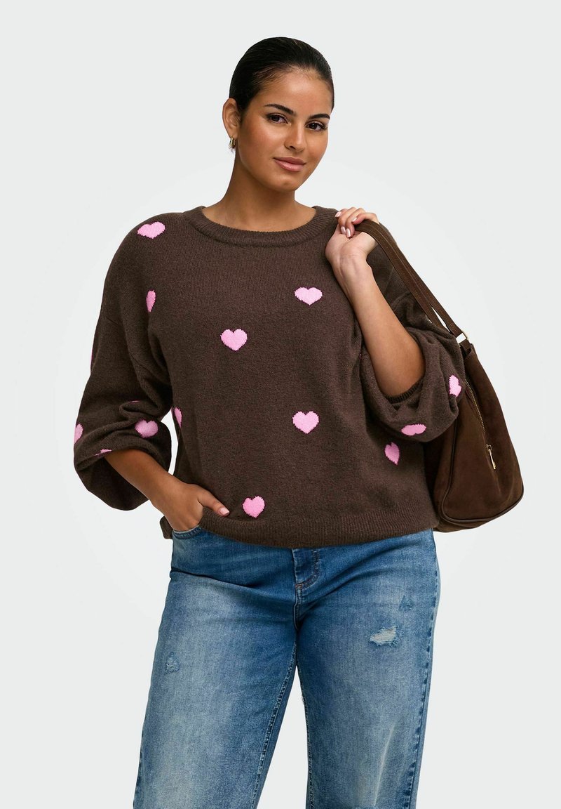 Femme portant un pull marron avec des cœurs roses, un jean bleu, et tenant un sac à épaule marron, sur un fond clair uni.