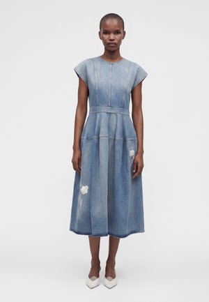 Kvinde iført en ærmeløs blå denim midi-kjole med hvid blomsterbroderi og hvide spidse hæle, står foran en ensfarvet baggrund.