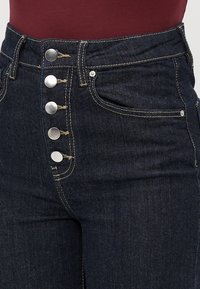 Mørkeblå høytlivsjeans med fem eksponerte sølvknapper og synlig beige søm, iført med en maroon topp.