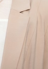 Blazer beige clair avec une texture lisse, muni de revers étroits et de détails de couture subtils. Le tissu semble léger et légèrement froissé.