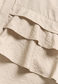 Beige layered fabric with three ruffled tiers, featuring a crinkled texture and clean stitching along the edges. Soft, lightweight material.