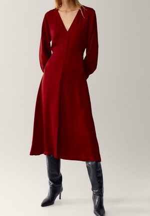Femme portant une robe midi à manches longues, de couleur rouge foncé avec un décolleté en V, associée à des bottes noires à talons et montantes jusqu'aux genoux, posant devant un fond uni.