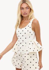 Femme blonde portant un débardeur blanc et un short à pois noirs, regardant sur le côté avec une expression neutre.