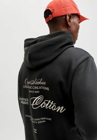 Must kapuutsiga dressipluus, millel on suur kapuuts ja valge trükitud tekst seljal, valmistatud puuvillast ja mugava lõikega.