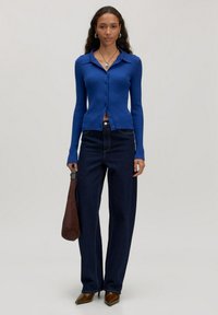 Blauwe geribbelde cardigan met knopen, gedragen over hoog-waisted donker denim jeans. Het model houdt een bruine handtas vast en staat op puntige bruine hakken.