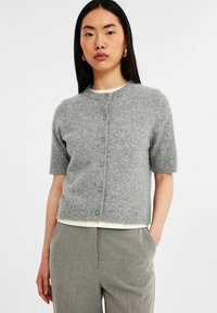 Grå stickad kortärmad cardigan med rund halsringning, knappfront och mjuk textur. Bärs över en vit topp, med grå byxor.